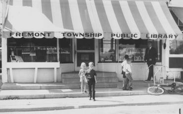 Original Fremont Township Library picture from the 1950s features two children standing in front of the building and two children and a gentleman walking past the front doors. The awning reads "Fremont Township Public Library".